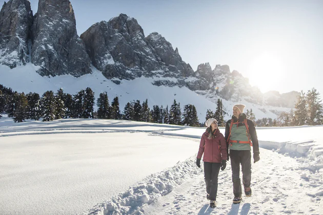Ein Mann und eine Frau wandern durch eine verschneite Winterlandschaft, hinter ihnen die Geislerspitzen