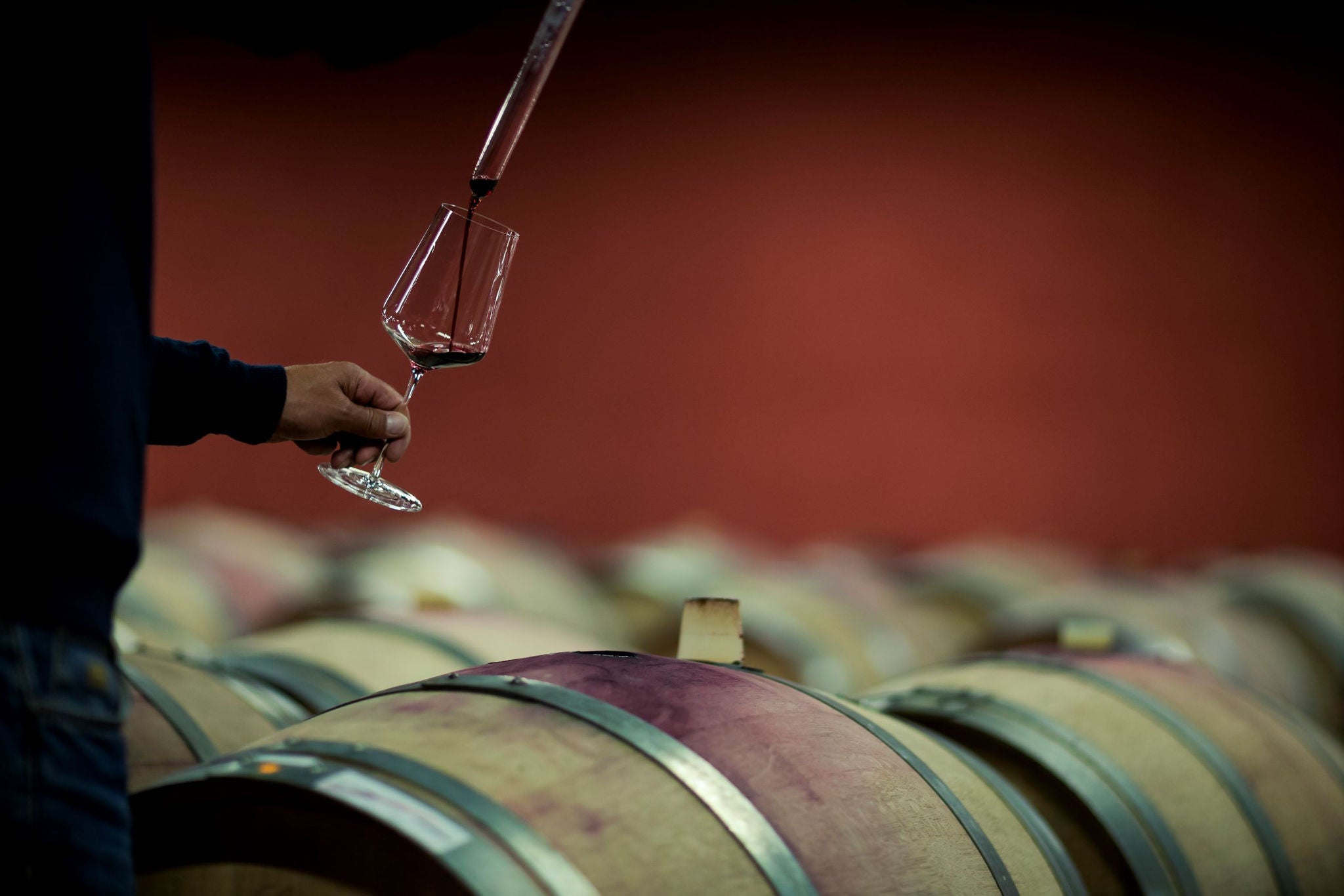 Ein Kellermeister steht vor einem Weinfass aus Hols und füllt über eine Pipette etwas Rotwein in ein Glas.