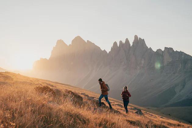 Zwei Personen wandern bei Sonnenaufgang über eine Bergwiese, im Hintergrund schroffe Felsen