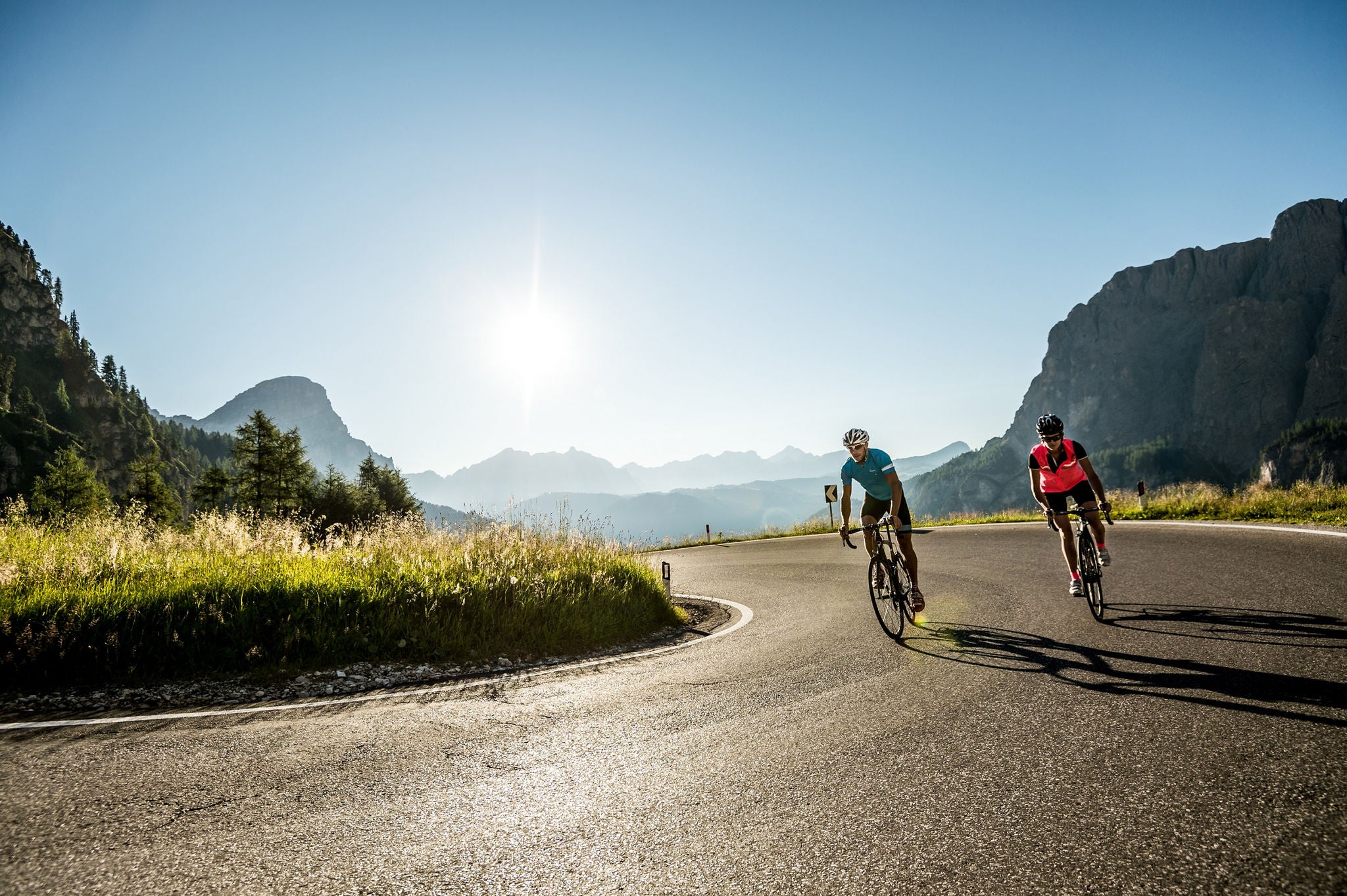 Mit der Sonne im Rücken fahren ein Mann und eine Frauf auf ihren Rennrädern einen Bergpass in Alta Badia hinab.