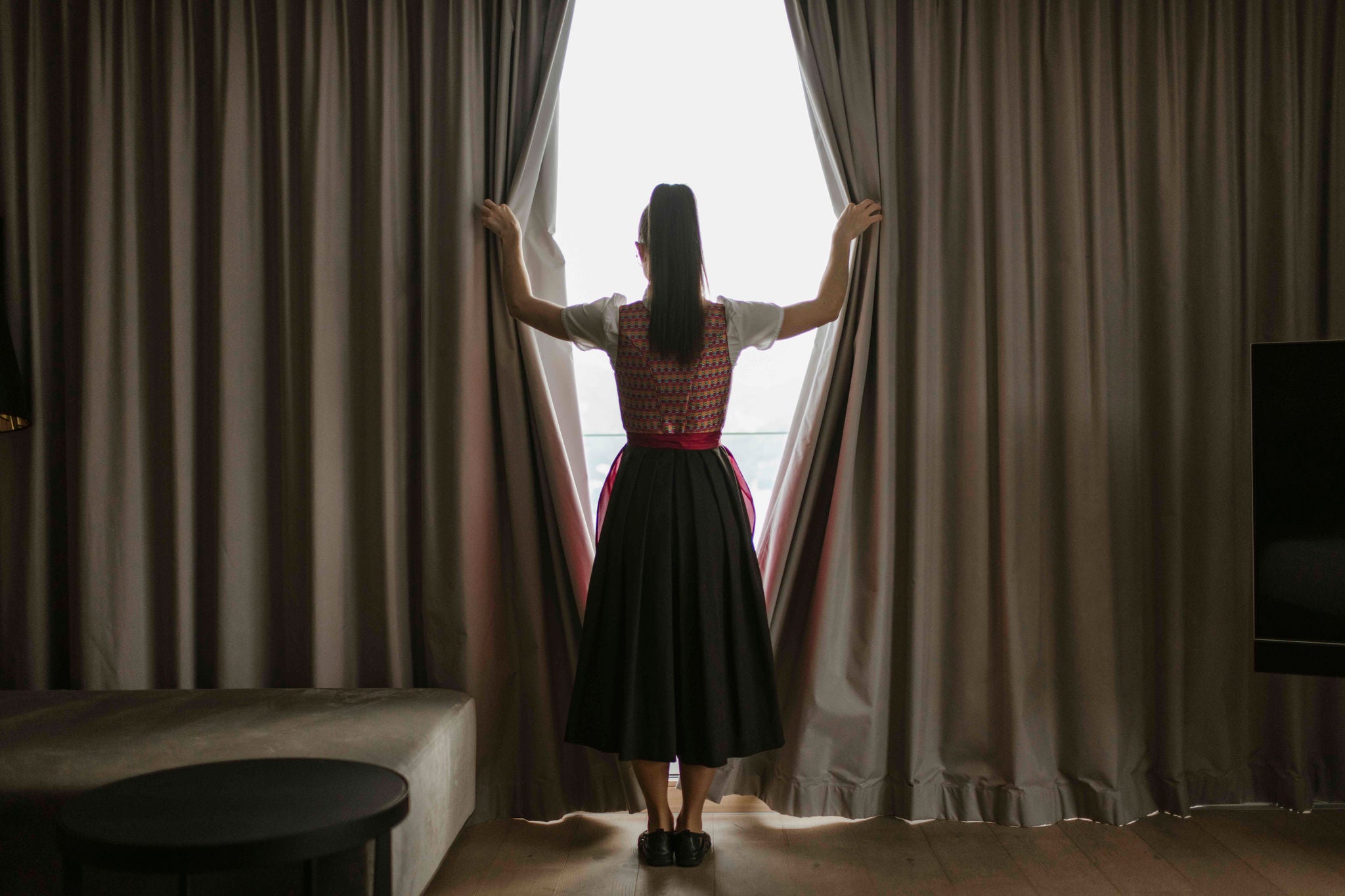 Eine Frau in traditioneller Tracht steht an einem Fenster und zieht die Vorhänge auf