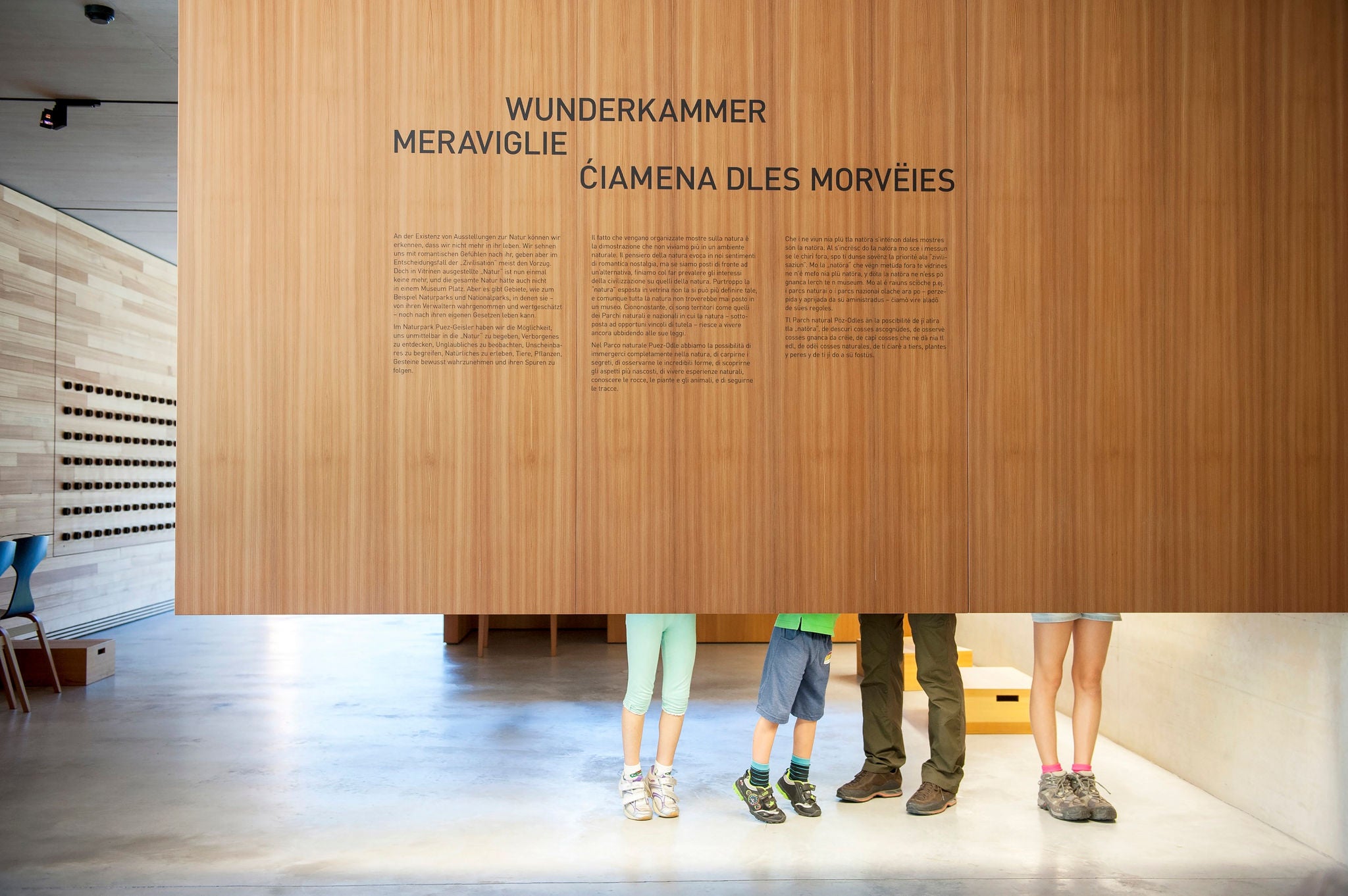 Die Füße von vier Besuchern des Naturparkhauses Villnöss sind hinter einer Ausstellungswand mit allen drei Südtiroler Sprachen Italienisch, Deutsch und Ladinisch.