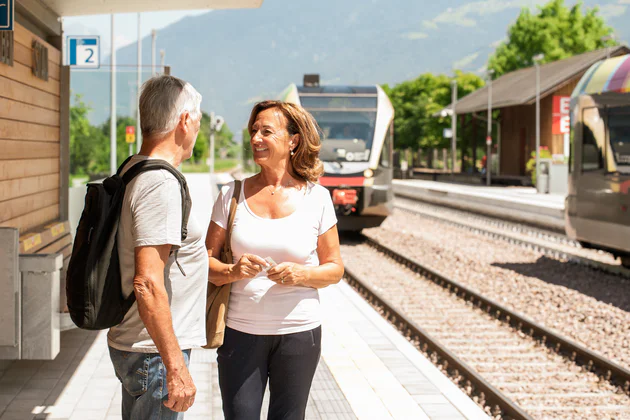 A man and woman standing at the station, she is smiling at him. A train can be seen in the background.