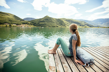Eine Frau mit langen Haaren sitzt in Wanderklamotten und barfuß am Steg des Kalterer Sees