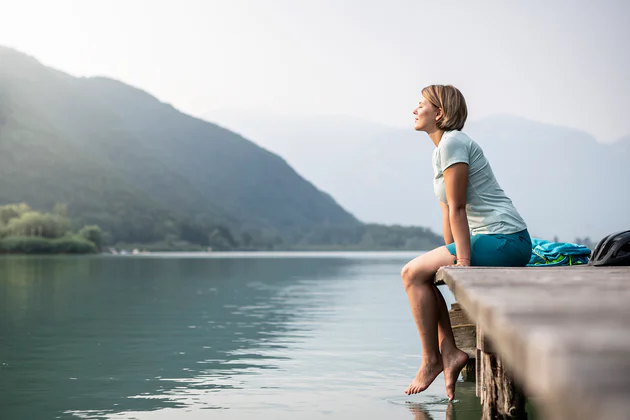 Eine Frau mit kinnlangen blonden Haaren sitzt mit geschlossenen Augen am Steg des Kalterer Sees