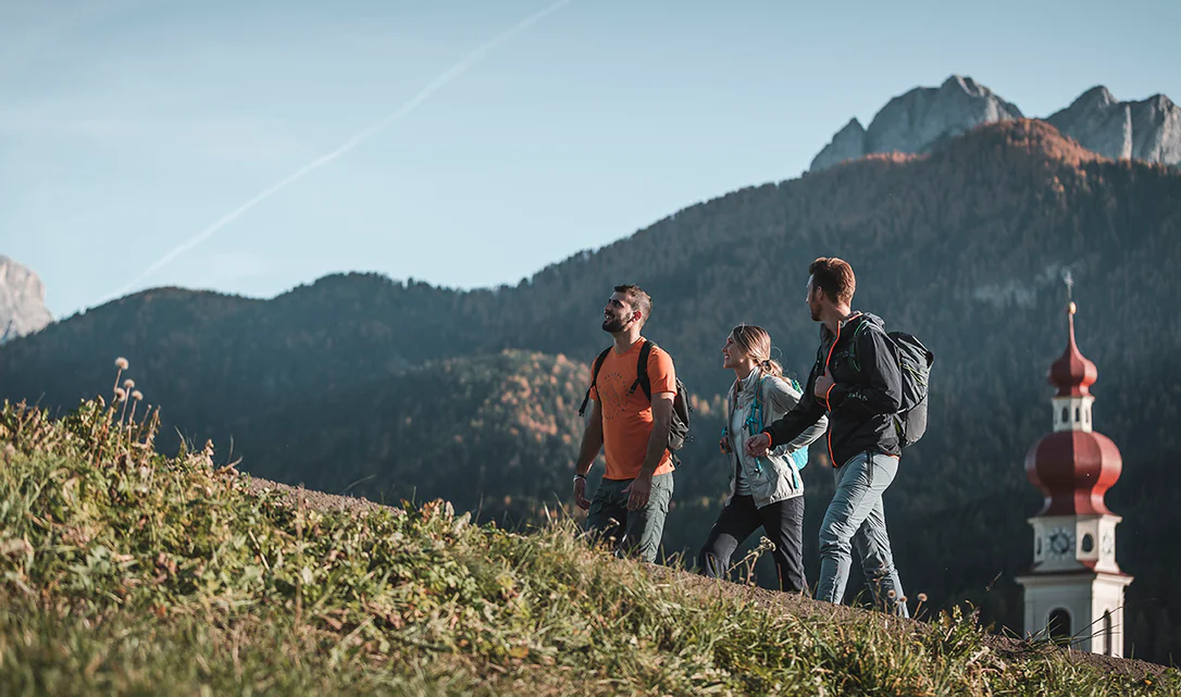 3 Personen wandern in Südtirol, im Hintergrund ist ein krichturm mit roter Spitze zu sehen