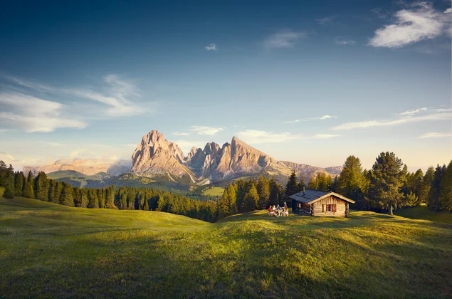 Úchvatný pohled na zelenou horskou louku s dřevěnou chatou, v pozadí jsou sluncem ozářené horské štíty