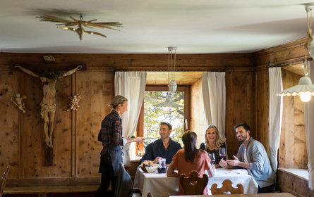 In einer traditionellen Holzstube sitzen 4 Gäste an einem Tisch und werden von einer blonden Kellnerin mit karierter Bluse bedient.