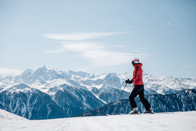 Een persoon met een rode jas staat op een helling op ski's en kijkt uit op een prachtige, besneeuwde bergtop