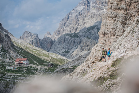 A man and a woman walking on a path alongside the famous 3 Zinnen Dolomites with a panoramic view on the "Dreizinnenhütte" mountain refuge.