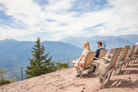 Due persone ammirano il panorama montano dall'alto, sedute su delle seggiole in legno