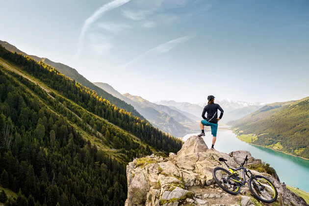 Osoba patrzy na góry w regionie Vinschgau, za nią na skale leży oparty rower