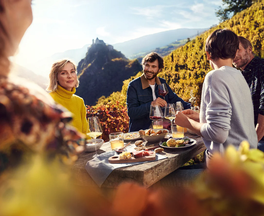 Fünf Leute genießen bei sonnigem Herbstwetter mit Wein und einer Brotzeit das Törggelen im Eisacktal. Im Hintergrund ist das frühere Kloster Säben zu erkennen.