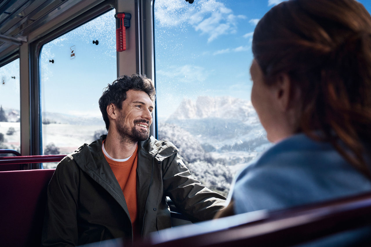 Ein junger Mann sitzt im Zug einer jungen Frau gegenüber, vor dem Fenster zieht die verschneite Bergwelt vorbei.