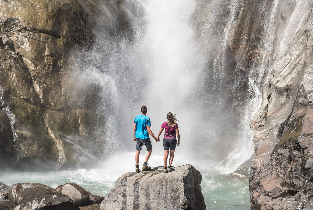 Zwei Person stehen vor einem Wasserfall und bestaunen das Naturschauspiel