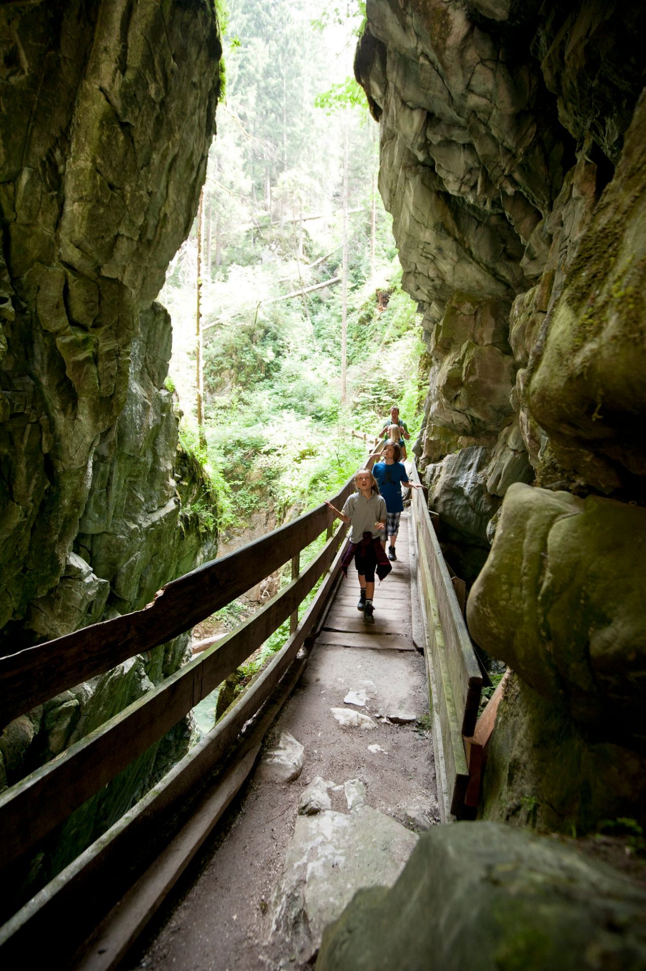 Eine Familie geht über eine schmale Holzbrücke, links und rechts von ihr ragen steile Felswände empor.