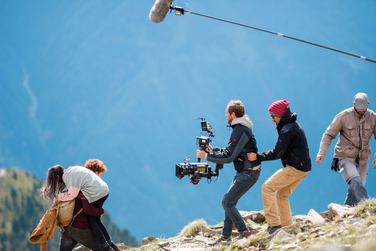 Ein Kamerateam dreht auf einem felsigen Abhang eine Filmszene, die einen jungen Mann zeigt, der ein junges Mädchen den Berg hinauf trägt.