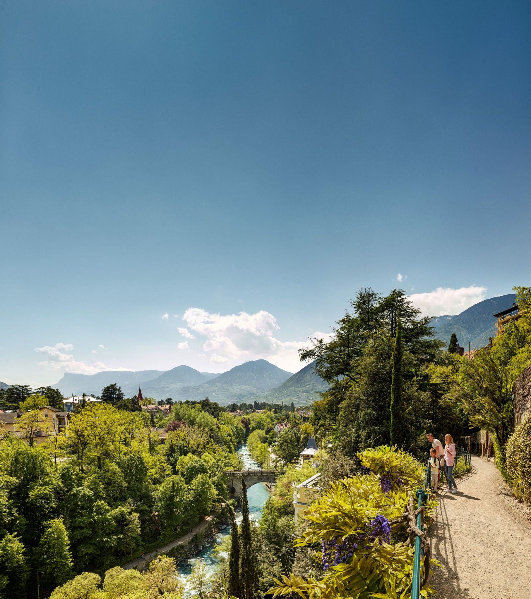 Familie auf einem sonnenverwöhnten Spazierweg mit Blick auf zahlreiche Bäume und einen Fluss, der durch Meran fließt.