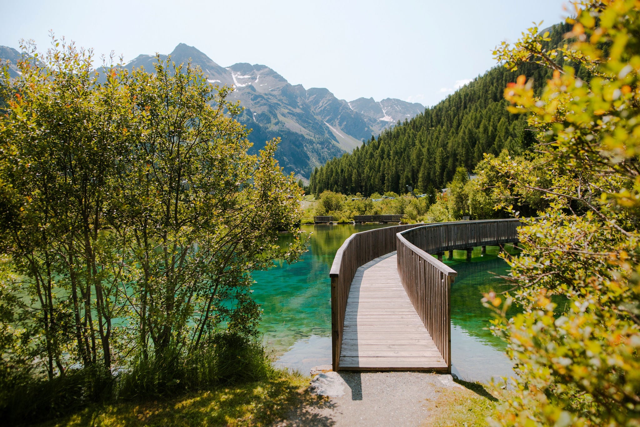 Ein Holzsteg führt über türkisgrünes Wasser des Suldensees, im Hintergrund der Ortler