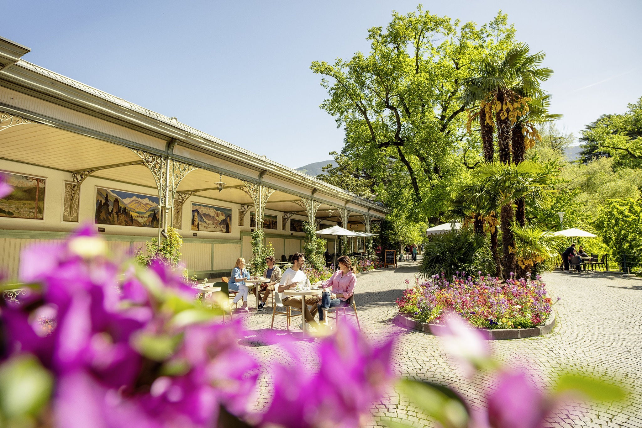 Vier Personen sitzen an Kaffeetischen vor der Wandelhalle in Meran an der von Blumenbeeten gesäumten Gilfpromenade 