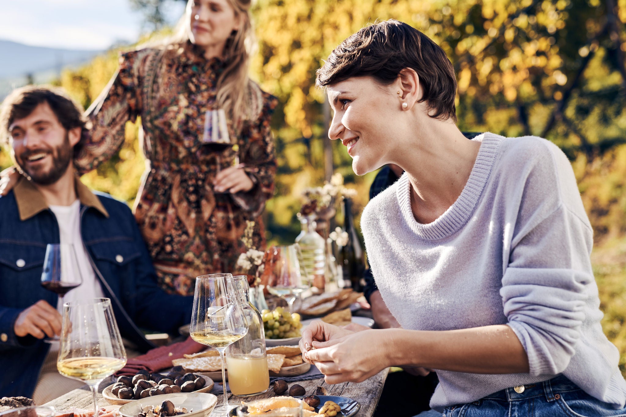 Eine lächelnde Frau an einer Törggelen-Tafel mit Kastanien, Wein, Speck und Trauben.