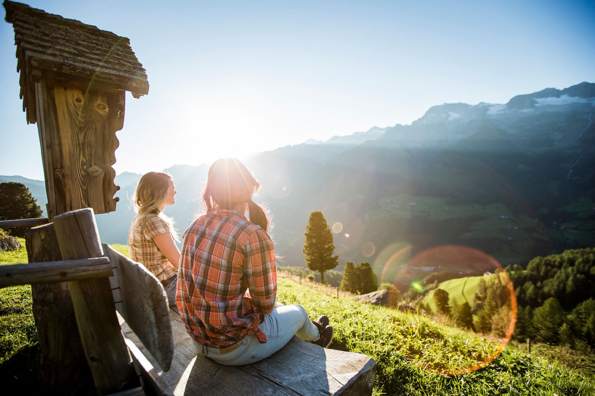 Zwei Frauen rasten auf einer Bank vor einem Wegkreuz und genießen das Panorama