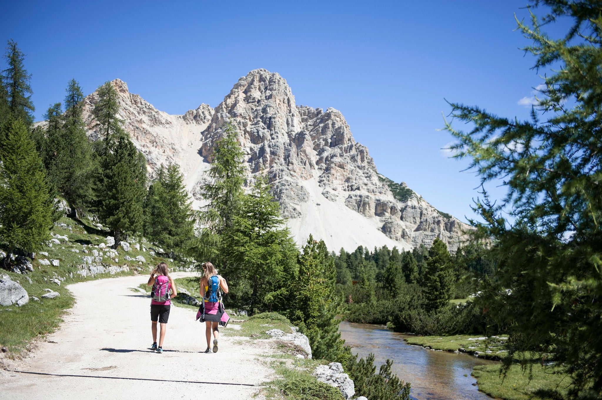 Zwei Frauen wandern auf einem Wanderweg, der neben einem kleinen Bach entlang läuft, in der Bergwelt der Dolomiten.