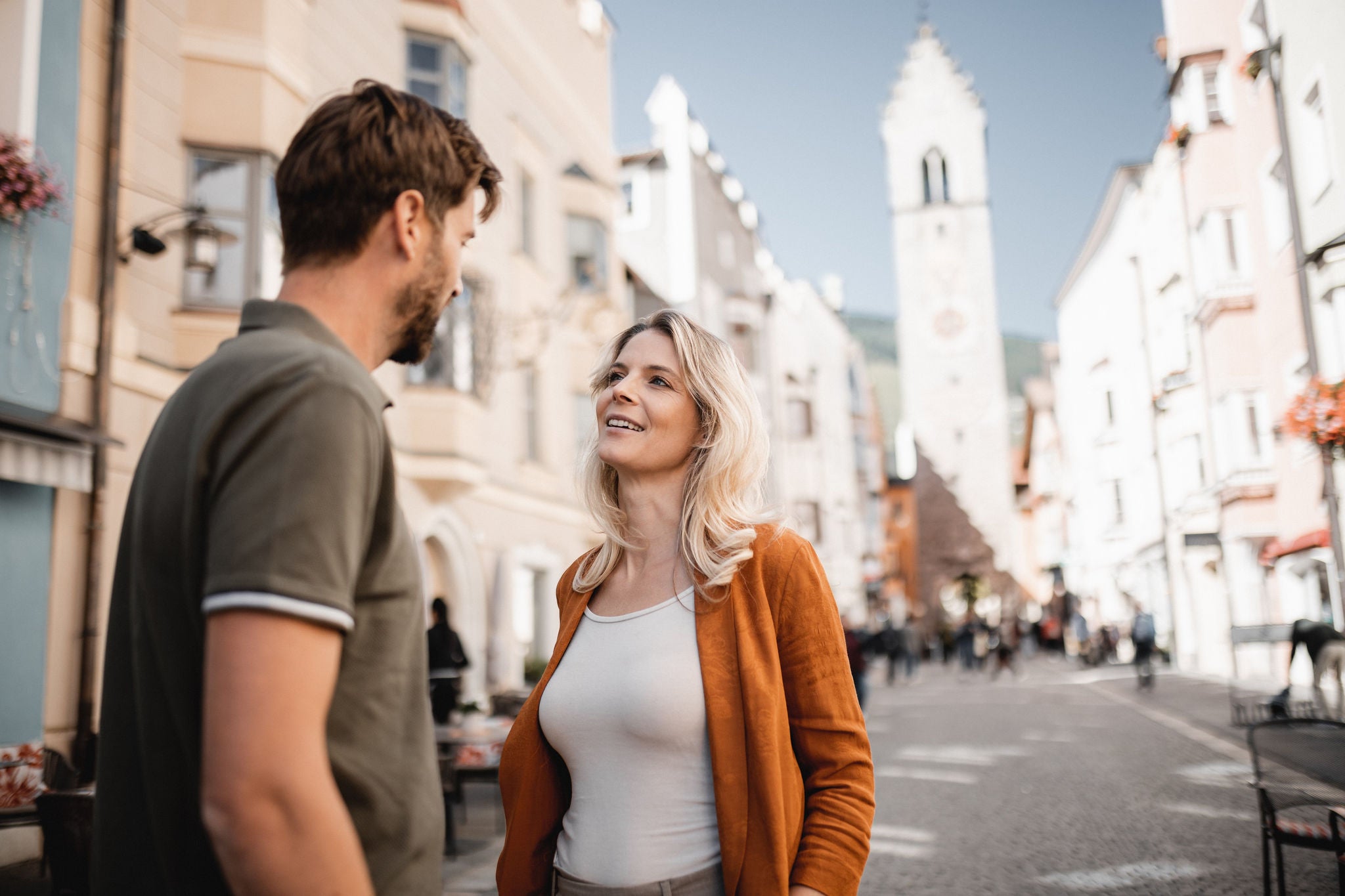 Eine Frau und ein Mann im Sommerlook sprechen miteinander in der Stadtgasse von Sterzing, die vom ZwÃ¶lferturm dominiert wird.