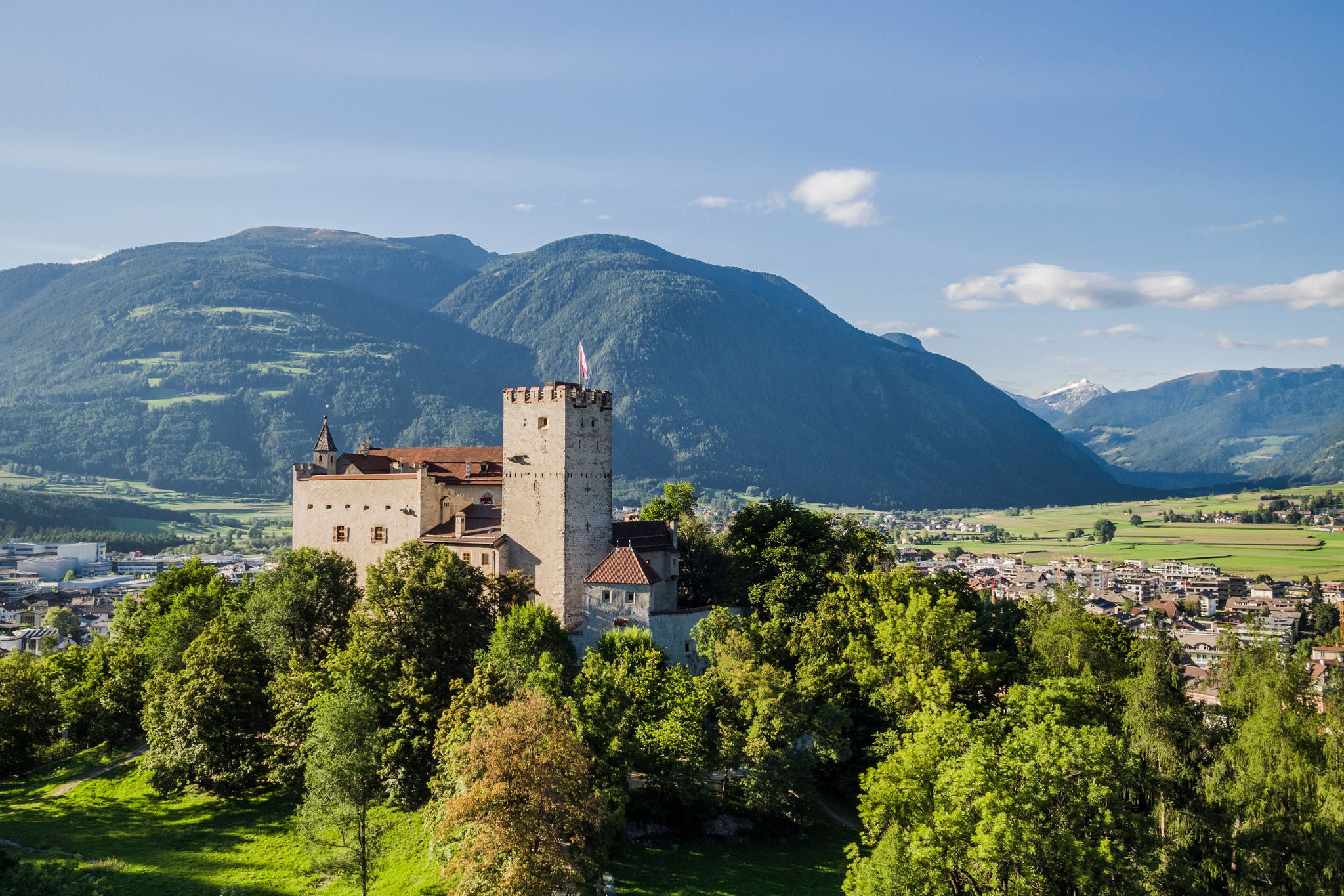 Vorne erhebt sich das Schloss Bruneck, dahinter erstrecken sich die Ausläufer der Stadt Bruneck und grüne Wiesen