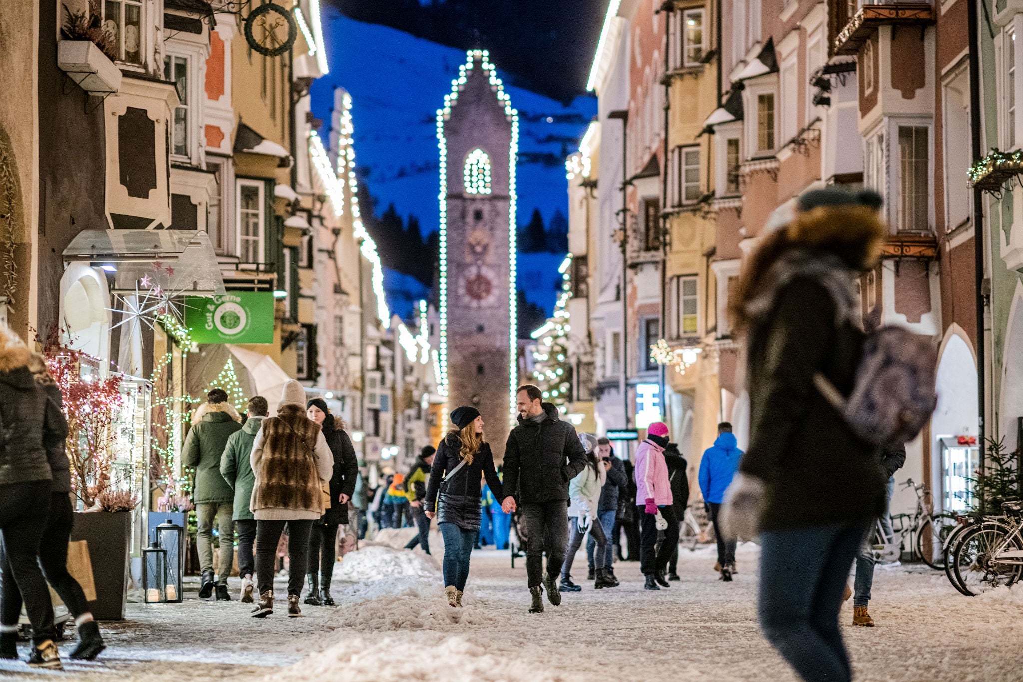 Die historische Stadtgasse von Sterzing mit beleuchtetem Zwölfertrum an einem Winterabend mit bummelnden Menschen.