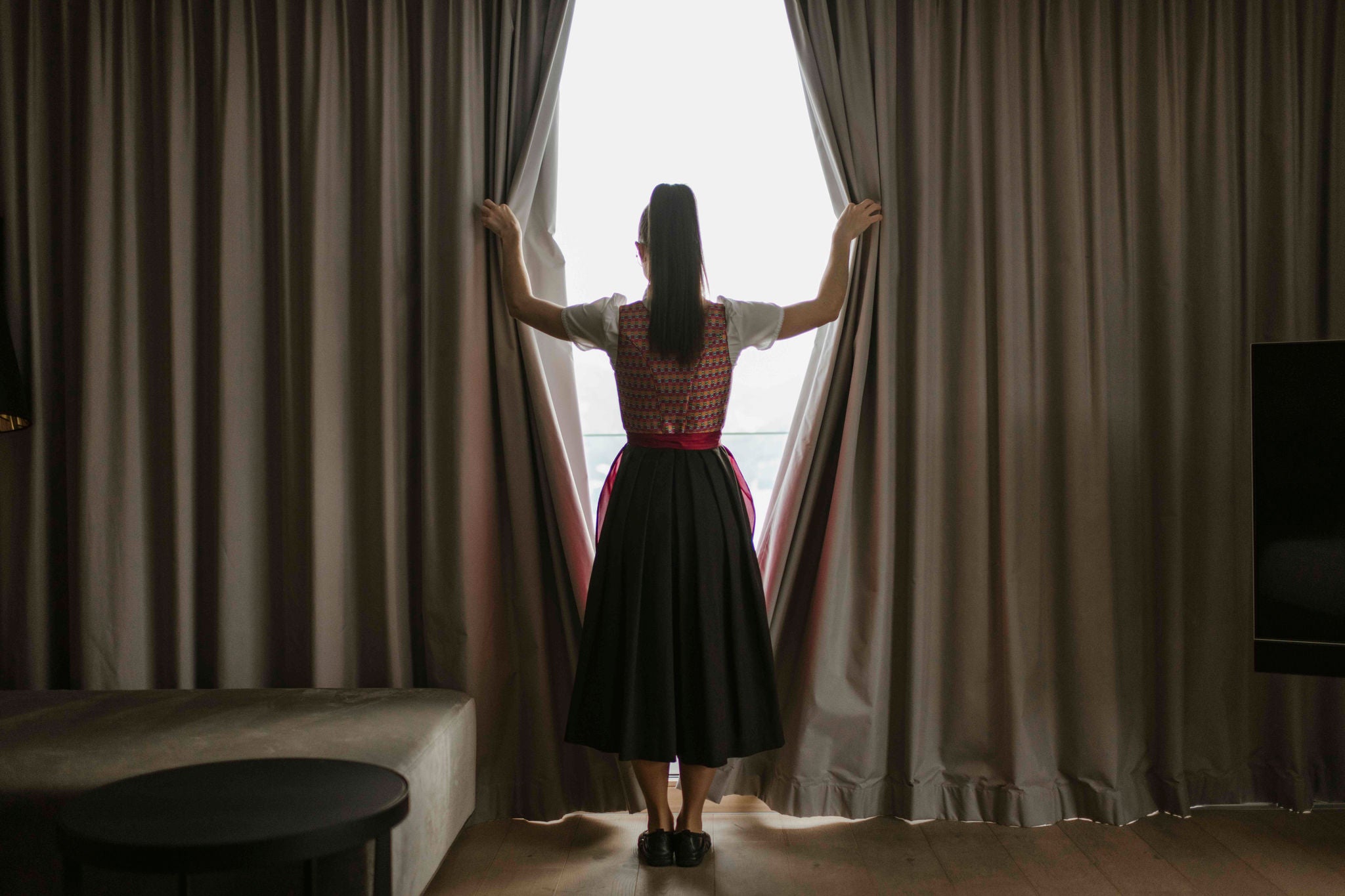 Eine Frau in traditioneller Tracht steht an einem Fenster und zieht die Vorhänge auf
