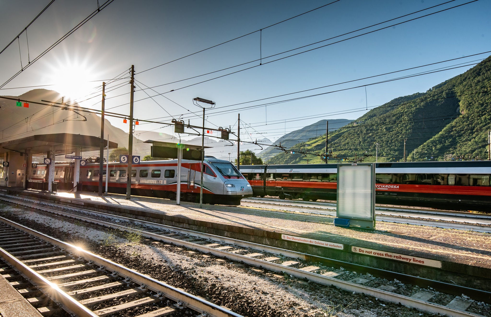 Zwei Züge in der Sonne im Bahnhof von Bozen mit der Bergkulisse im Hintergrund.
