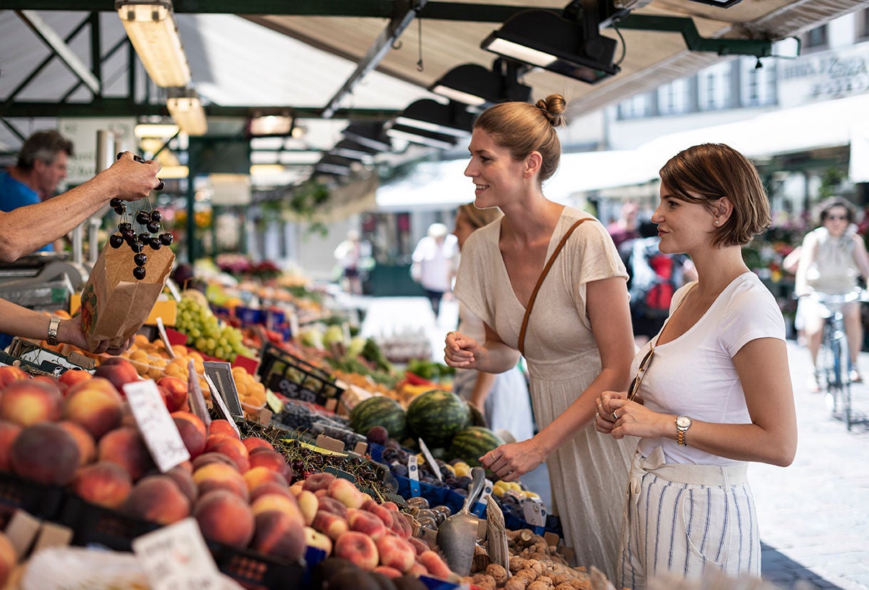 Zwei Personen lassen sich von einem Händler Obst auf dem Obstmarkt zeigen