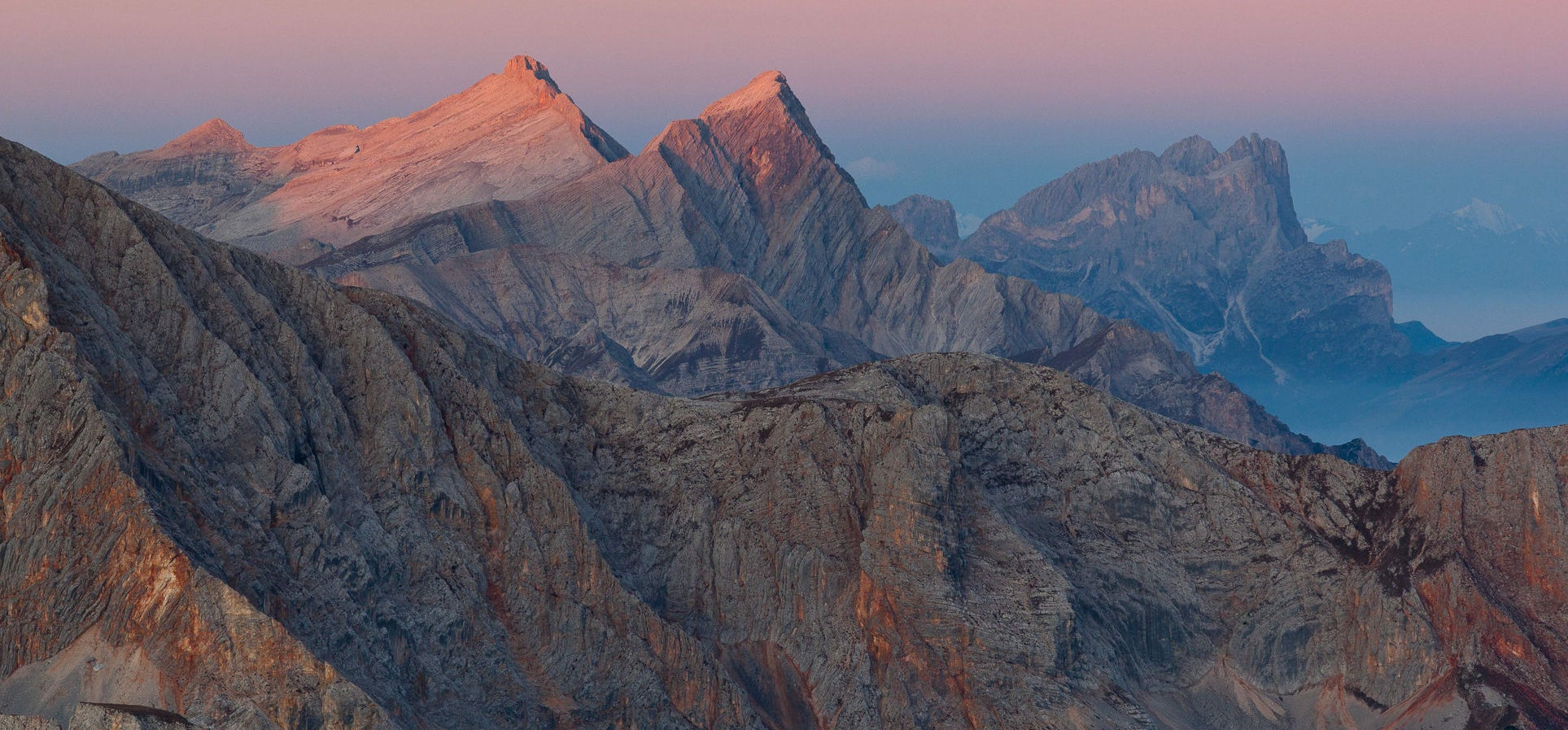 Gesteinsschichten unterschiedlicher Farbtöne ergeben einen massiven Berg, dahinter leuchten zwei Bergspitzen im Morgenrot.