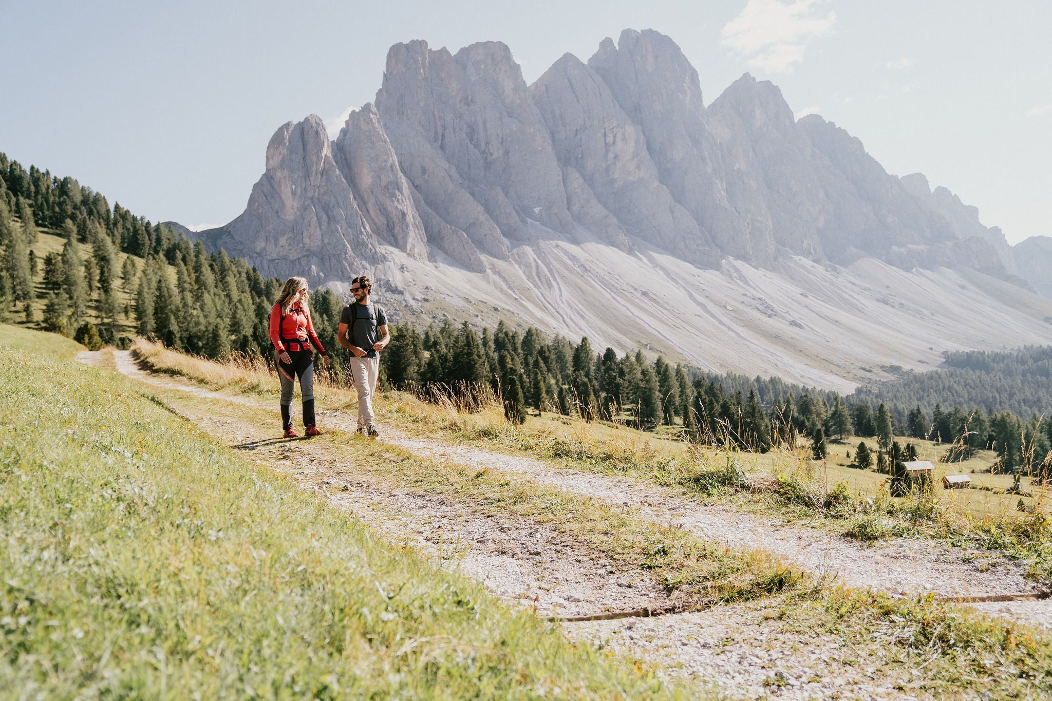 Ein Mann und eine Frau wandern über einen Schotterweg, dahinter erheben sich die Villnösser Geisler in den blauen Sommerhimmel.