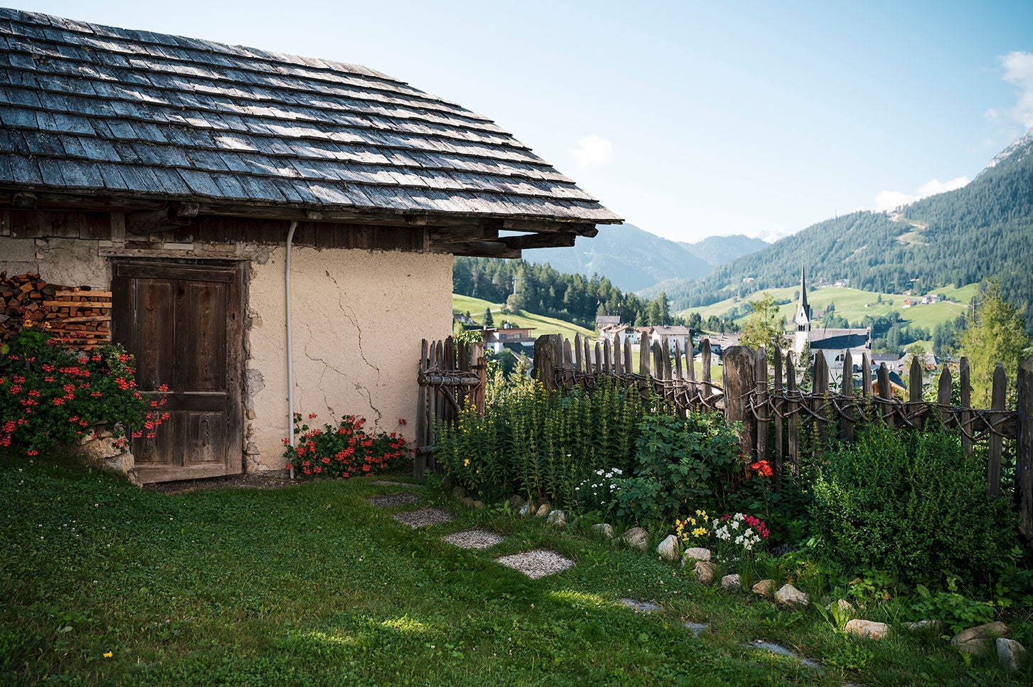 Sommerlicher Bauerngarten mit Kräutern, der zu einem alten Bauernhof mit Schindeldacht gehört, dahinter ist die Dorfkirche zu sehen