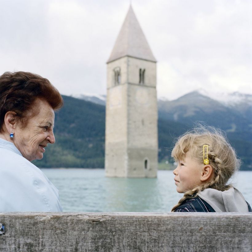 Die braunhaarige Theresa Theiner und ihre blonde Enkelin sitzen sich gegenüber am Ufer des Reschensees mit dem Kirchturm des früheren Ortes Graun.
