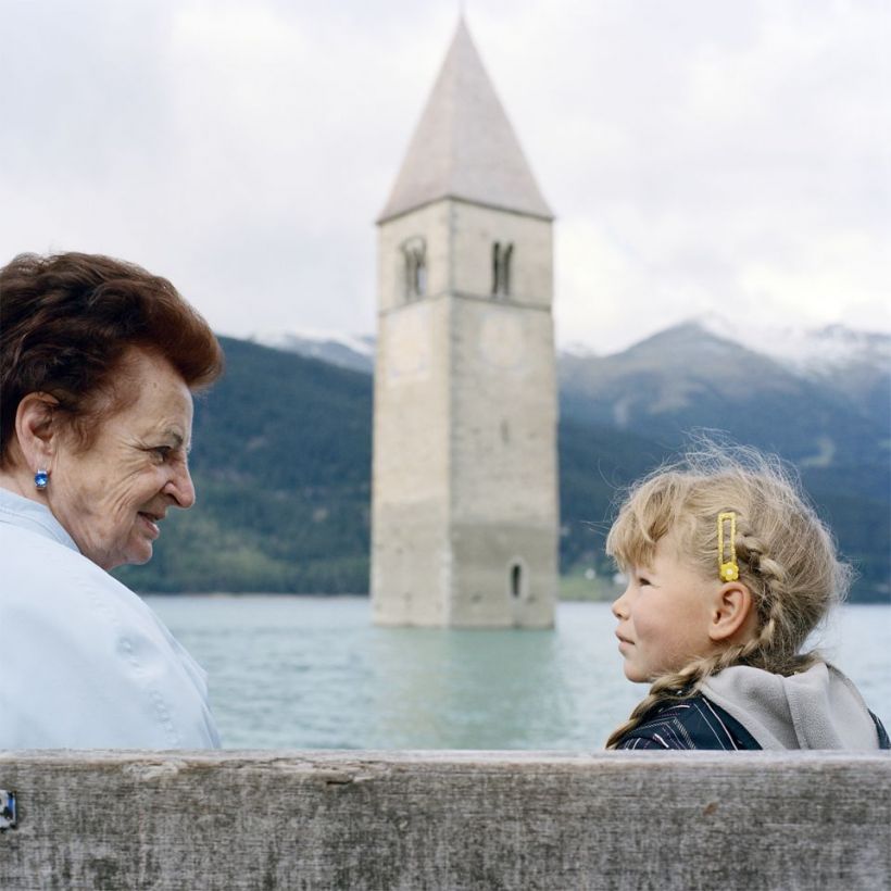 Die braunhaarige Theresa Theiner und ihre blonde Enkelin sitzen sich gegenÃ¼ber am Ufer des Reschensees mit dem Kirchturm des frÃ¼heren Ortes Graun.