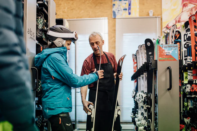 Een vrouw met een skihelm en een blauwe jas krijgt advies van een man in een skiverhuurwinkel