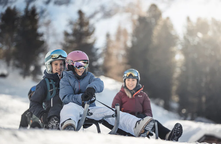 Two children on a toboggan going towards the valley. In the background an adult woman looking after them.