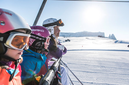 Twee kinderen en een vrouw in ski-uitrusting zitten op een stoeltjeslift met een besneeuwde piste op de achtergrond