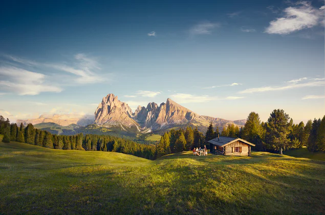 Atemberaubender Blick auf eine grüne Bergwiese mit Holzhütte, im Hintergrund von der Sonne beleuchtete Berggipfel