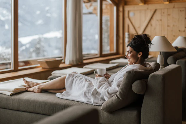 EIne Frau im Bademantel entspannt in einem Wellnesshotel. In der Hand hält sie eine Tasse und sie blickt aus der großen Fensterfront vor ihr