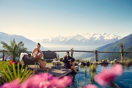 Deux femmes en train de profiter de vacances bien-être dans le Sud-Tyrol. Elles sont assises sur une terrasse au bord d'un étang de baignade, dans un jardin en fleurs. D'imposantes montagnes enneigées sont visibles en arrière-plan.