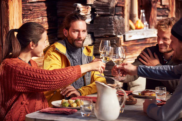 Groupe de 4 personnes (1 femme et 3 hommes) dans un chalet, assis autour d'une table et trinquant avec un verre de vin blanc à la main