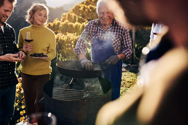 Several people with wine glasses watch a smiling man roasting chestnuts in a pan.