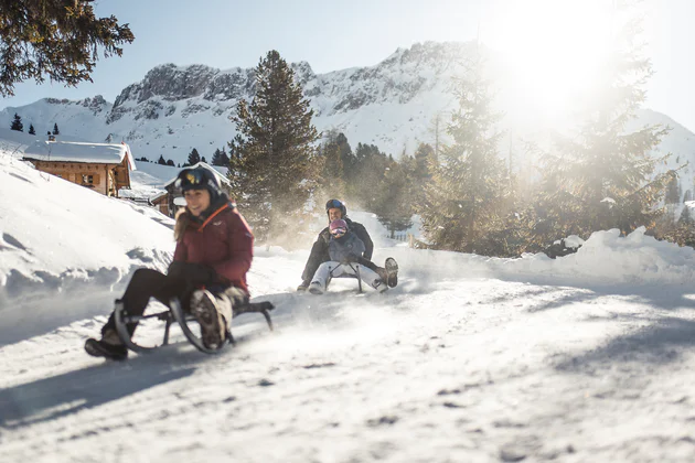 Una mamma e un papá con il bambino ricendono divertiti una pista innevata in slittino in Val di Funes