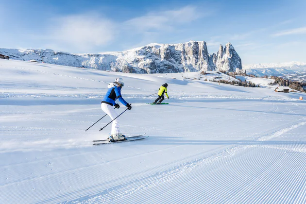Two skiers ski down the slope in front of a mountain panorama.