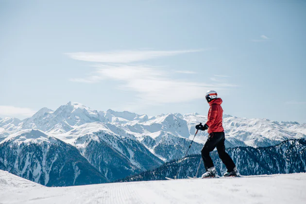 Skieur en veste rouge se tenant sur une piste de ski et contemplant un magnifique sommet enneigé