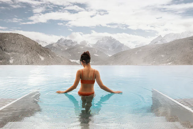 Femme se tenant debout dans une piscine en train de contempler les montagnes devant elle. Elle a de l'eau jusqu'à la taille et fait glisser ses mains sur la surface de l'eau.